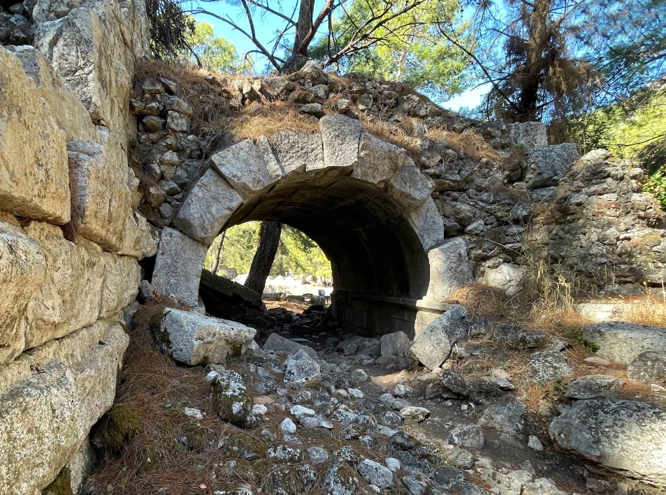 Ancient stone archway in a rocky ruin, with trees overhead and sunlight through the tunnel.