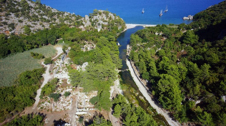 Aerial view of a green, rocky coastal landscape with a narrow waterway running between hills toward a sandy beach and blue sea with sailboats offshore.