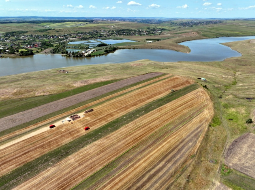 Aerial view of striped harvested fields on a hillside, with hay bales and red trailers, and a winding river with a village in the distance.