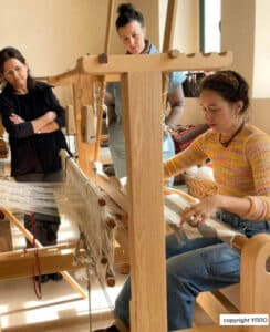Three women weave on a large wooden loom in a bright workshop setting with natural light.