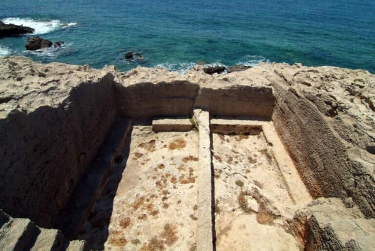 Rocky coastline with a sunken rectangular stone trough or pool carved into the rock, overlooking blue sea.
