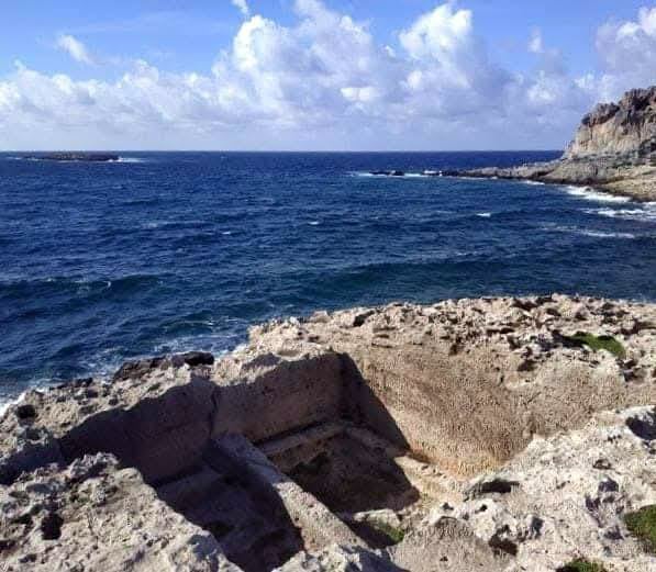 Rugged rocky coastline with blue ocean and a clear sky, waves lapping against the rocks.
