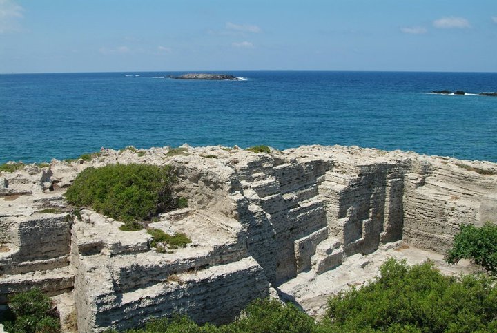 Coastline with layered limestone rock formations and sparse green shrubs overlooking a blue sea under a clear sky