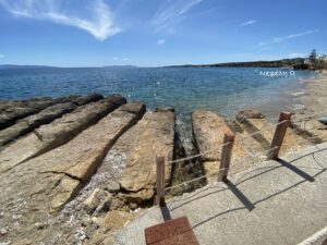 Coastal scene with rocky breakwater, clear blue sea, and a paved walkway with rope railing along the shore under a bright sky.