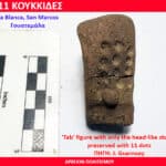 Brown clay 'Tab' figurine fragment with a head-like stump and 11 dot impressions, shown beside a measurement scale (inches and cm).