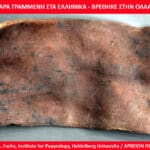 Ancient copper tablet with scratched Greek inscription, dark patina and irregular edges on a gray surface.