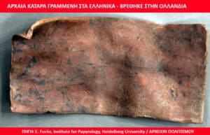 Ancient copper tablet with scratched Greek inscription, dark patina and irregular edges on a gray surface.