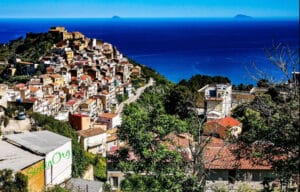 Colorful hillside town clinging to the coast, with a dense cluster of pastel houses and a winding road leading down toward the sea; deep blue Mediterranean backdrop.