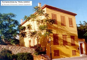 Δύο-όροφο κίτρινο σπίτι με καφέ παντζούρια, δέντρα μπροστά, και πετρόχτινος τοίχος κατά μήκος του δρόμου.