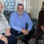 Three adults sit on a patterned couch in a casual indoor setting, looking toward the camera and smiling slightly. Peripheral items and a window with a metal grid are in the background.