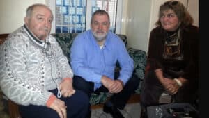 Three adults sit on a patterned couch in a casual indoor setting, looking toward the camera and smiling slightly. Peripheral items and a window with a metal grid are in the background.