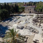 Ancient ruin site with scattered stone blocks, columns, and a stone building in the background, surrounded by trees.