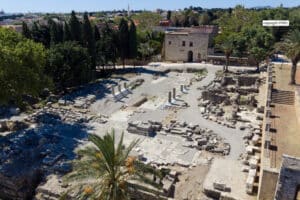 Ancient ruin site with scattered stone blocks, columns, and a stone building in the background, surrounded by trees.