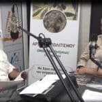 Two men sit across from each other in a radio studio, speaking into large microphones with papers and magazines on the desk behind a banner with Greek text.