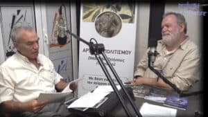 Two men sit across from each other in a radio studio, speaking into large microphones with papers and magazines on the desk behind a banner with Greek text.