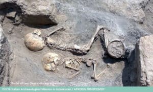 Two ancient child skeletons buried in a shallow grave in Uzbekistan, with a clay vessel nearby and surrounding soil.