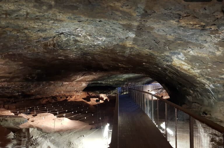 Wooden boardwalk with a metal railing winds through a dim underground cave with rugged rock ceiling and walls.