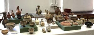 Assortment of ancient ceramic vessels, jugs, bowls, and plates displayed on green platforms in a museum exhibit.