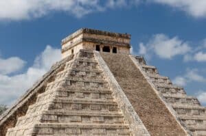 Stone stepped pyramid with a rectangular temple on top against a blue sky with clouds, resembling a Mesoamerican ruin