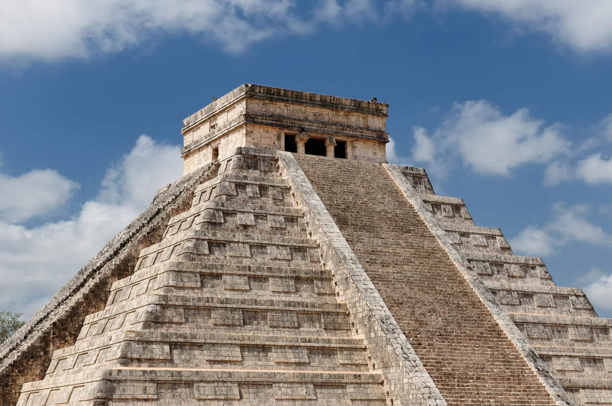 Stone stepped pyramid with a rectangular temple on top against a blue sky with clouds, resembling a Mesoamerican ruin