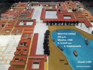 Scale model of an ancient city with red-roofed buildings surrounding a large blue pool; Greek caption and 1:300 scale noted on the model.