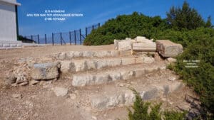 Ancient stone steps and scattered blocks along a dirt path with green bushes and a blue sky; Greek photo credit in corner.