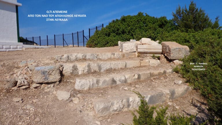 Ancient stone steps and scattered blocks along a dirt path with green bushes and a blue sky; Greek photo credit in corner.