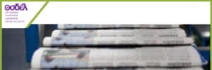 Green-framed banner showing a Greek organization logo in the top-left and a close-up of stacked newspapers on a printing press in the rest of the image.