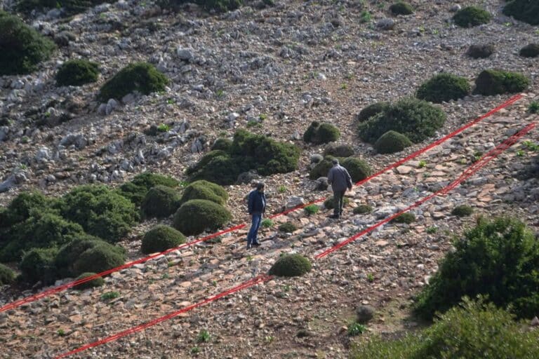 Two hikers walk along a rocky hillside marked by red tape lines amid low shrubs and stones.