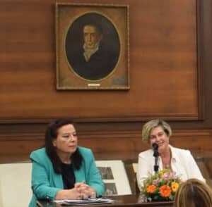 Two women seated at a panel table in a room with wood-paneled walls; one in turquoise blazer, the other in white, speaking into a microphone with a flower arrangement nearby.