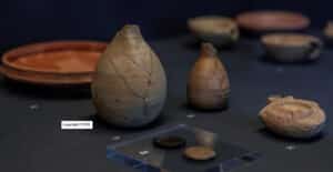 Collection of ancient clay vessels and coins displayed on a dark museum table with numbered markers