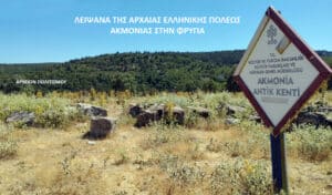 Rural archaeological site with dry grass, scattered stones, and a tilted sign reading 'AKMONIA ANTİK KENTİ' against a blue sky.