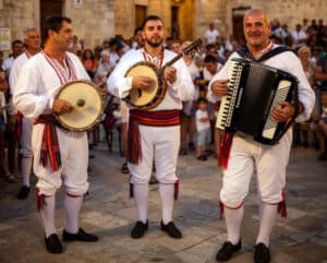 Three male musicians in white traditional outfits perform folk music in a sunlit town square, playing tambourines/mandolin and an accordion while a crowd watches.