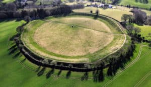 Aerial view of a circular earthwork or hill fort with a grassy central area and a surrounding bank, trees along the rim and fields beyond.