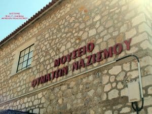 Stone museum exterior with large red Greek lettering on the wall announcing the Nazi Crimes Museum (exposed stone façade).