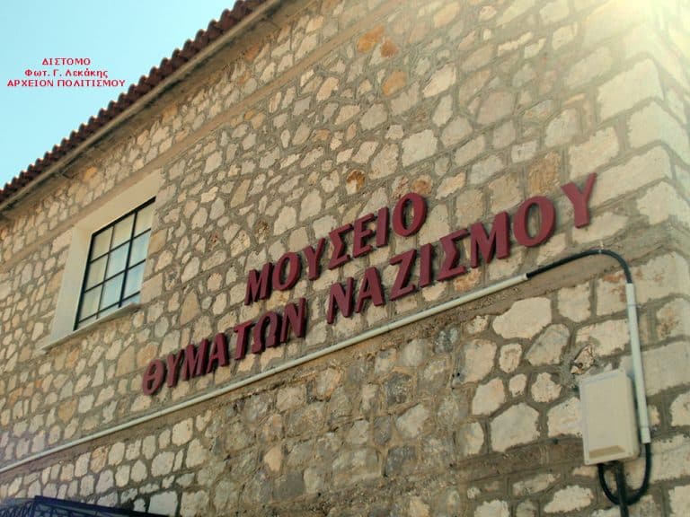 Stone museum exterior with large red Greek lettering on the wall announcing the Nazi Crimes Museum (exposed stone façade).