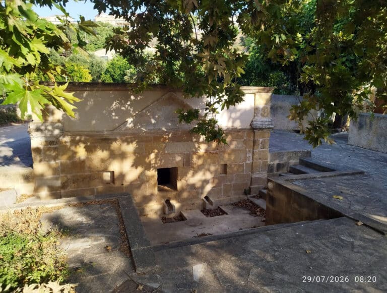 Stone brick structure in a garden courtyard with a triangular pediment and small square openings, shaded by overhanging leaves.