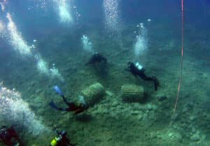 Scuba divers explore a rocky seabed with large concrete blocks.
