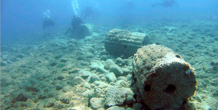 Underwater scene of a rocky seabed with large, rounded rocks and divers in the distance swimming above.
