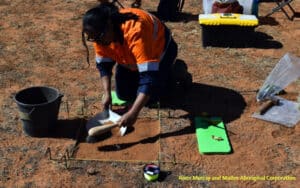 Individual in an orange safety vest kneels at a grid-outlined dirt patch, carefully scraping soil with a trowel for archaeological sampling.