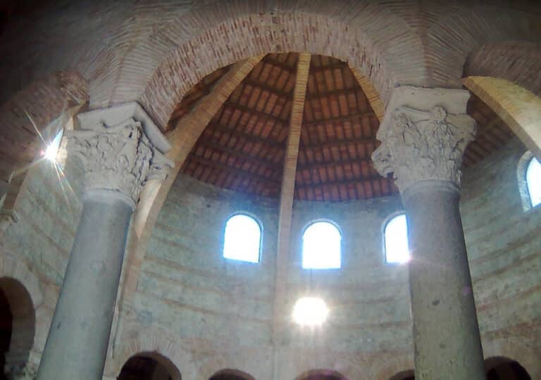 Inside a circular stone hall with tall columns and arched windows under a wood-beamed dome roof, sunlight streaming in