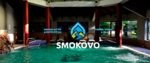 Indoor spa pool area with red columns and a large Smokovo logo over the water; signs read 'THERMAL SPRINGS & SPA' and Greek text on either side.
