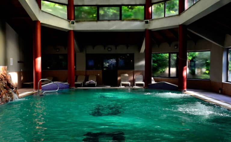 Indoor pool with turquoise water, red columns, lounge chairs along the back wall, and large windows showing greenery outside.