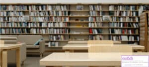 Quiet library interior with light wood tables and chairs, and tall book shelves filled with books in the background.