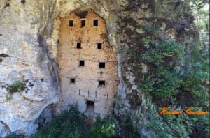 Ancient multi-story cliff dwelling carved into a rock wall, with many small square windows and surrounding greenery; watermark 'Kostas Gerlos' in the lower right corner.