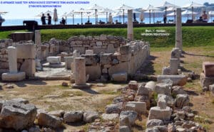 Seaside archaeological site with ancient stone walls and columns, and visitors under umbrellas by the shore in the background.