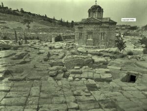 Ruined stone courtyard with large rectangular blocks, a small brick church in the distance on a hilltop.