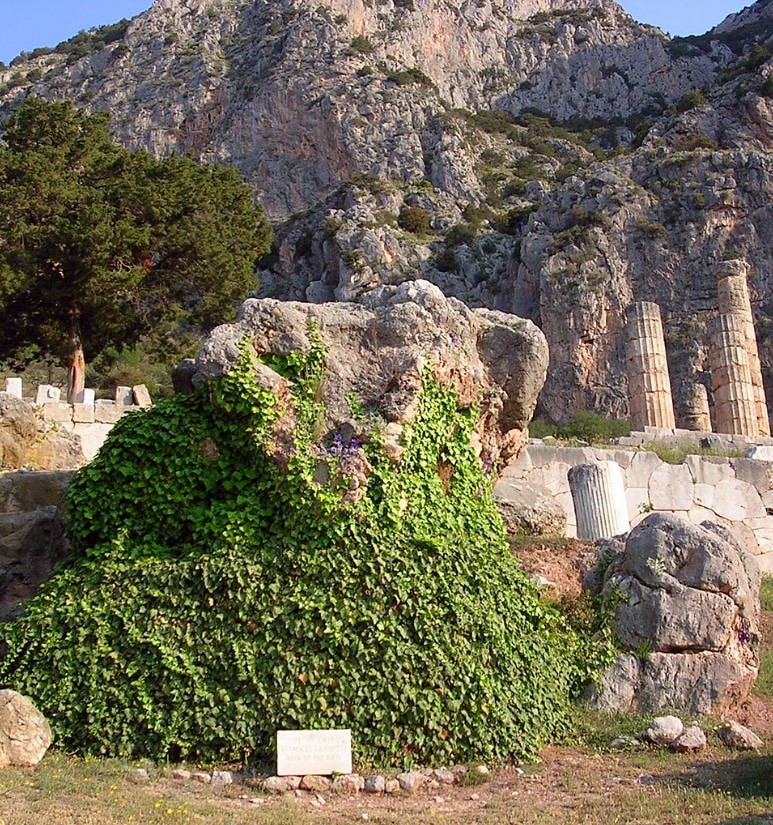 Ivy-covered ancient statue or ruin in front of rocky hills with broken column fragments nearby.