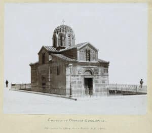 Small stone church with a domed roof and decorative façade, fenced in, on a bare hill with two distant figures nearby.