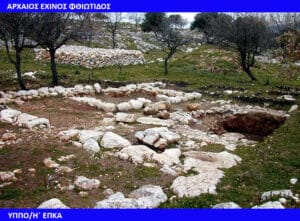 Ancient stone walls forming terraces on a grassy hillside with olive trees; an excavation trench in the foreground.